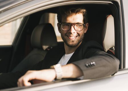 Attractive young man with a beard and glasses smiles while looking out the window of his car.