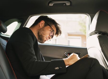 Young and attractive man with a beard and glasses works writing on some documents inside a car. Businessの写真素材