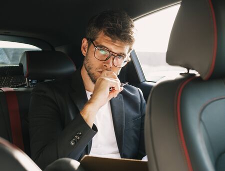Young and attractive man with a beard and glasses works and looks at camera inside a car. Businessの写真素材