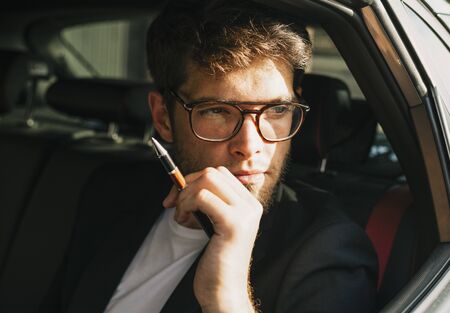 Attractive young man with a beard and glasses looks through the window of his car. Businessman.の写真素材