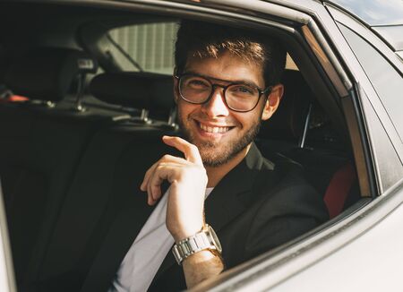 Attractive young man with a beard and glasses smiles while looking through the window of his car. Businessman.の写真素材