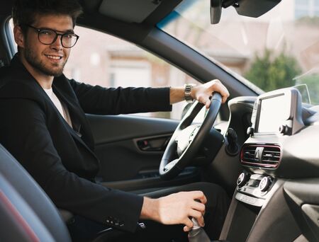 Attractive young man with a beard and glasses smiles at the camera in his car.の写真素材