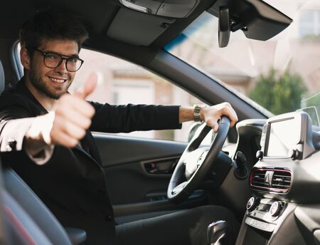 Attractive young man with a beard and glasses smiles and says ok to the camera in his car.の写真素材