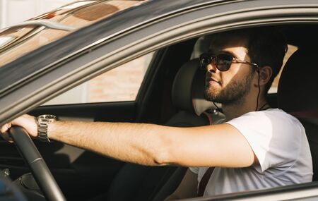 Attractive young man with a beard and sunglasses drives his car.の写真素材