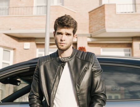 Attractive young man with a beard looks seriously at the camera while leaning on his carの写真素材