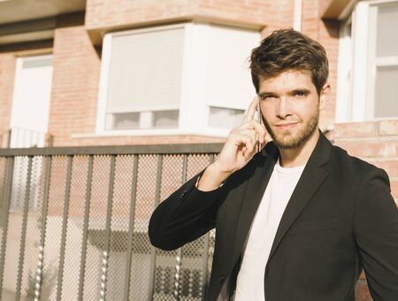 Close-up of attractive young man with beard in suit talks on the phone and smiles while looking at camera.の写真素材