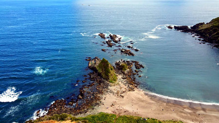 Aerial view of a rugged Pacific coastline with a sandy cove, tombolo-like rock spur, and scattered sea stacks. Clear blue water, gentle surf, and tide-washed reefs create a serene, natural seascape ideal for travel, nature, and coastal themes.の写真素材