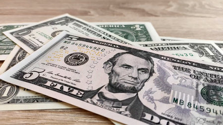 A crisp macro of U.S. currency on a wooden tabletop, featuring a five dollar bill in sharp focus with soft bokeh background. Perfect for finance, banking, savings, budgeting, investment, and economy concepts in ads, blogs, and presentations.の写真素材