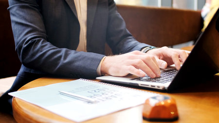 Close-up of a professional typing on a laptop at a wooden cafe table. Warm natural light, notebook and pen in frame. Ideal for themes of remote work, business, productivity, freelancing, and coworking.の写真素材