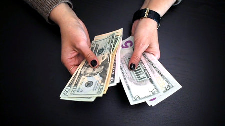 Close-up of female hands counting U.S. dollar bills on a dark desk, illustrating budgeting, savings,...