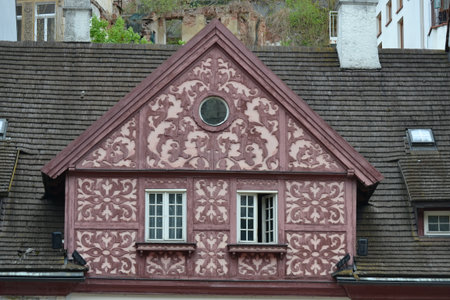 A detailed shot of a building's roof and gable, featuring a red and white patterned facade, it has two windows and a small round window, the weathered roof tiles and chimneys are also visibleの写真素材
