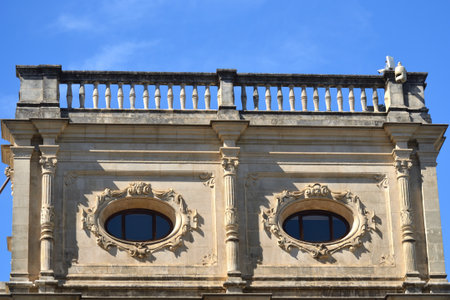 A low-angle view of a stone building with a balustrade. The facade features two identical oval windows framed by intricate scrolls and carvings, with fluted columns between them. A clear blue sky is visible above.の写真素材