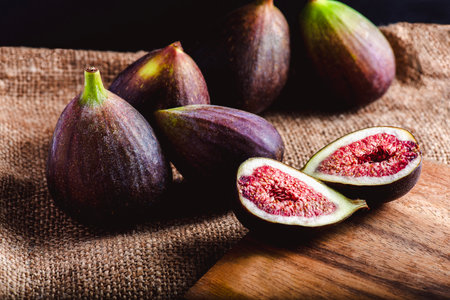 Ripe figs displayed on a rustic burlap cloth, with one fig cut open to reveal its vibrant red flesh and seeds, resting on a wooden surface under soft natural light.の写真素材
