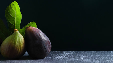 minimalist composition showcasing two ripe figs alongside a vibrant green fig leaf, set against a dark background with a textured surface for an earthy and natural feel.の写真素材
