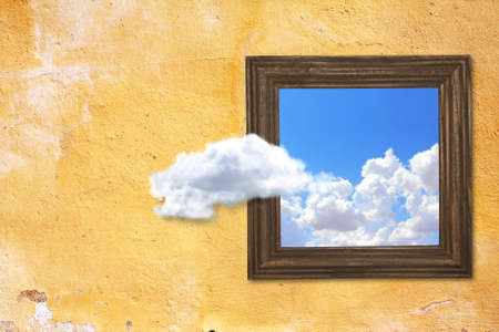 Think outside the box concept. Wooden picture frame with photo of blue sky with clouds at stucco wall background. A cloud floats out of a painting hanging on a wallの写真素材