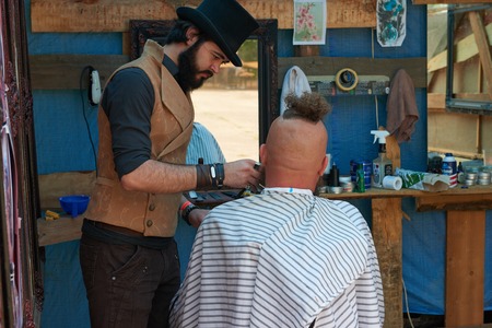 CHERNIVTSI, UKRAINE - JULY 22, 2017: Stylish hairdresser in the cylinder cap Shears and shaves a biker with red hair at the festival Moto-picnic Retrodrom 2017のeditorial素材