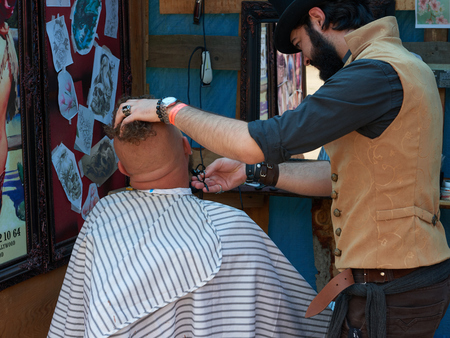 CHERNIVTSI, UKRAINE - JULY 22, 2017: Stylish hairdresser in the cylinder cap Shears and shaves a biker with red hair at the festival Moto-picnic Retrodrom 2017のeditorial素材