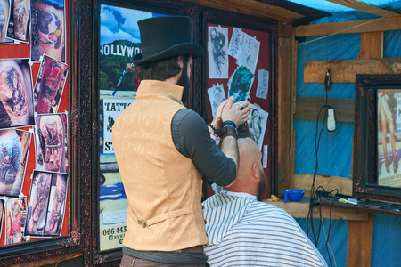 CHERNIVTSI, UKRAINE - JULY 22, 2017: Stylish hairdresser in the cylinder cap Shears and shaves a biker with red hair at the festival Moto-picnic Retrodrom 2017のeditorial素材