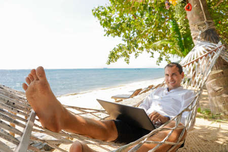 Young man work on laptop in white shirt relaxing in hammock with seaview. Remote workplaceの写真素材