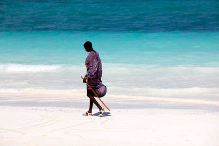 Zanzibar, Tanzania - January 5, 2016: Young massai man with colorful traditional clothes, headdress and self made shoes walking through the water on the Nungwi beach.のeditorial素材