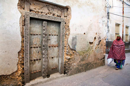 Stone Town, Tanzania - January 1, 2016: Woman in traditional Muslim clothes walking on the street of Stone Town. On her left side there is traditional house with old door and in background is another house.のeditorial素材