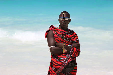 Zanzibar, Tanzania - January 5, 2016: Young massai man with colorful traditional clothes, headdress and self made shoes walking through the water on the Nungwi beach.のeditorial素材