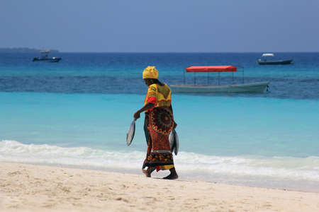 Zanzibar, Tanzania - January 7, 2016: An African woman in traditional dress with fish walks down the beach. Zanzibar, Tanzaniaのeditorial素材