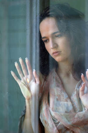 Woman with sad smile behind a wet windowの写真素材