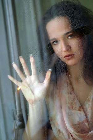 Woman with sad smile behind a wet windowの写真素材