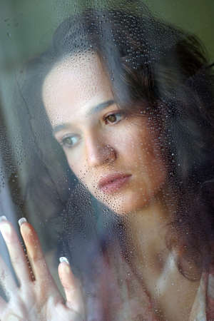 Woman with sad smile behind a wet windowの写真素材