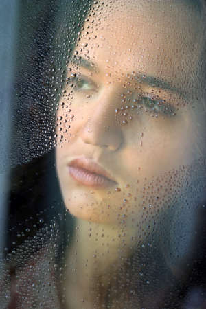 Woman with sad smile behind a wet windowの写真素材
