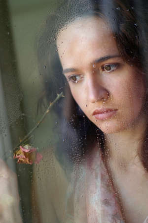 Woman with sad smile behind a wet windowの写真素材