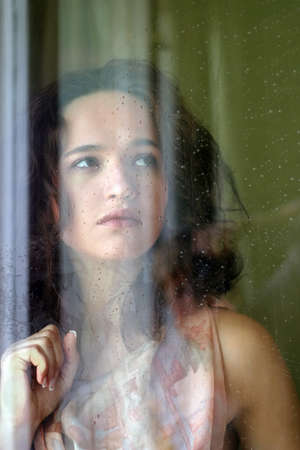Woman with sad smile behind a wet windowの写真素材