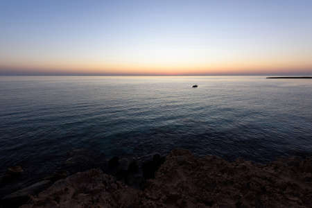 Sunrise with reflection of light in the water, in protaras paralimni, a fishing boat, dark blue sea and rocks, cyprus islandの写真素材
