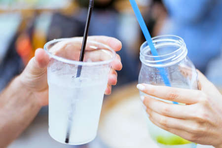 Drinking lemonade cocktail glasses with straw and ice shaking, man and woman friend hands cheers at an event with blurred red hair people and colored wall backgroundの写真素材