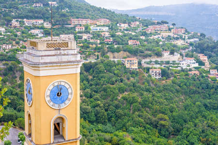 Daylight foggy view to Our Lady of the Assumption Church Clock Tower in Eze Village, French Riviera. Medieval Houses and green trees on background.の写真素材