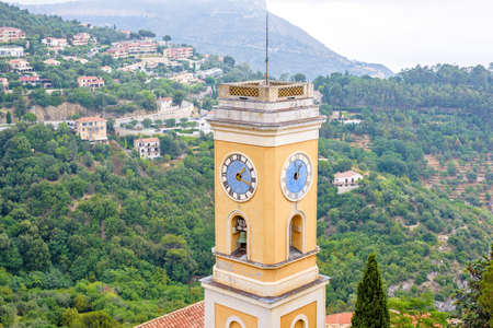 Daylight foggy view to Our Lady of the Assumption Church Clock Tower in Eze Village, French Riviera. Medieval Houses and green trees on background.の写真素材