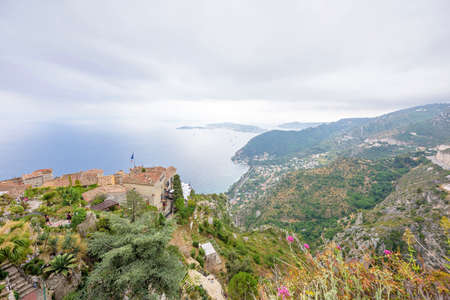 Scenic view from top to Mediterranean sea and part of Eze village. Medieval houses with France flag and green mountains in a foggy day of a French Rivieraの写真素材