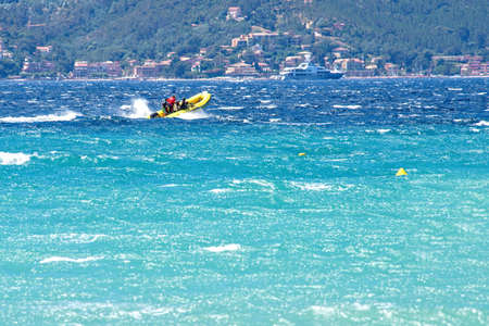 Daylight view to boat riding on water with people on it. Buoys, yacht, buildings and big green mountain on background. Cannes, Franceの写真素材