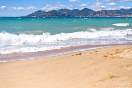 Daylight sunny view to blue sea and sand with traces from water. Focus on sand. Relaxing waves, green mountains and bright sky with clouds. Negative copy space. Place for text. Cannes, France.の写真素材