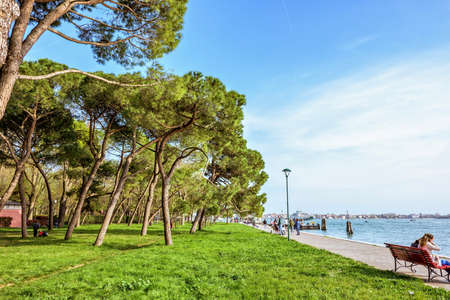 VENICE, ITALY - APRIL 02, 2017: Daylight view to vibrant green sunny park with tourists laying on grass,walking on alley and sitting on bench. City buildings and bright blue sky with cloudsのeditorial素材