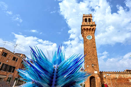 Daylight view to colorful Blue Murano Glass Sculpture with historic architecture clock tower on background. Bright blue sky with clouds. Murano Island, Venice, Italyの写真素材