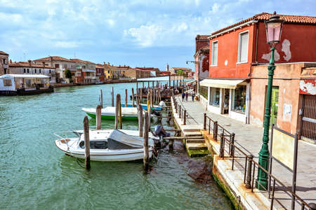 Daylight view to Venetian Lagoon and parked boats. People walking on sidewalk near colorful historic architecture buildings. Murano Island, Venice, Italyの写真素材