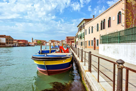 Daylight view to Venetian Lagoon canal with colorful parked boats and tourists walking on sidewalk. Bright blue sky with clouds and historic architecture buildings. Murano Island, Venice, Italyの写真素材