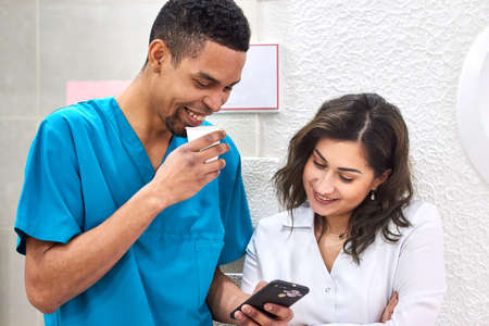 Young male african and female caucasian dentists on coffee break. Smiling and showing stuff on the smartphoneの写真素材