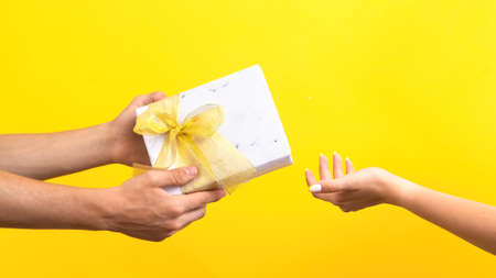 Man giving a gift box with red tape to a woman on yellow background. Hands. Love concept.の写真素材