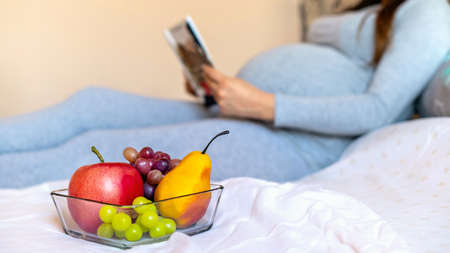 Pregnant woman lying on the bed and reading, dish with fruits on the foreground