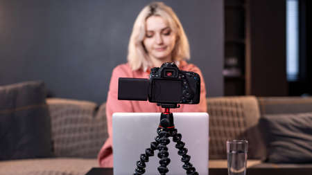 Young content creator blonde girl is on her laptop on the table. Filming herself using a camera on a...