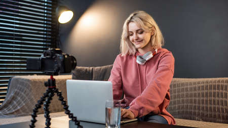 Young content creator smiling blonde girl with headphones working on her laptop on the table with camera on a tripod, microphone. Working from homeの写真素材