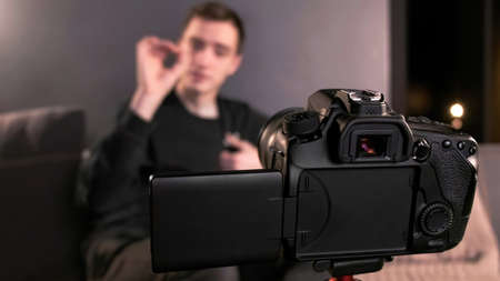 Young content creator talking and gesturing man filming himself using a camera on a tripod while...